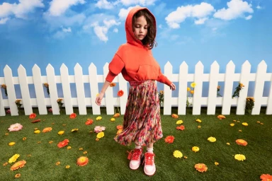 Child model wearing an orange hoodie and patterned pleated skirt in a garden set with white fence
