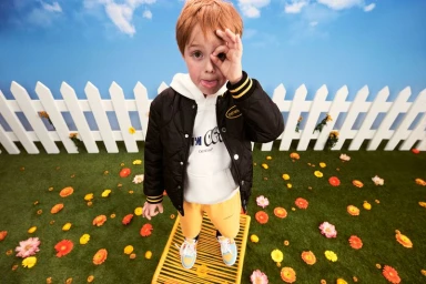 Young child in black jacket and white hoodie making ok sign with fingers over eye against white fence and blue sky backdrop
