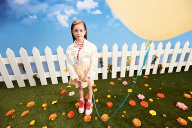 Young child wearing a white denim jacket and skirt standing on artificial grass holding a blue balloon string.