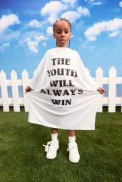 Young child holding up an oversized white t-shirt with black printed text against a backdrop of artificial grass, white picket fence, and a blue sky.