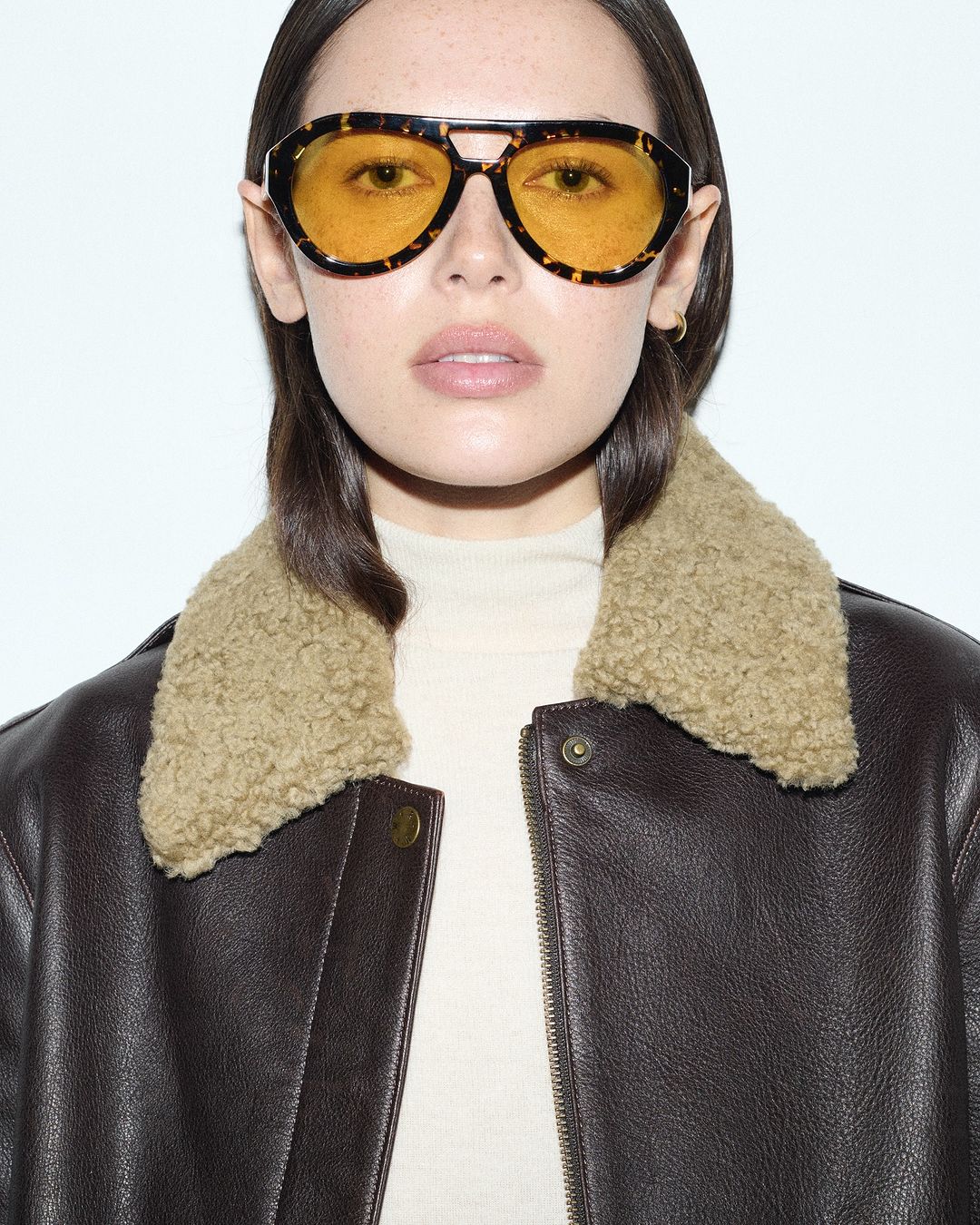 Portrait of a brunette woman wearing a dark leather jacket with a shearling collar and yellow-tinted sunglasses in a studio setting.
