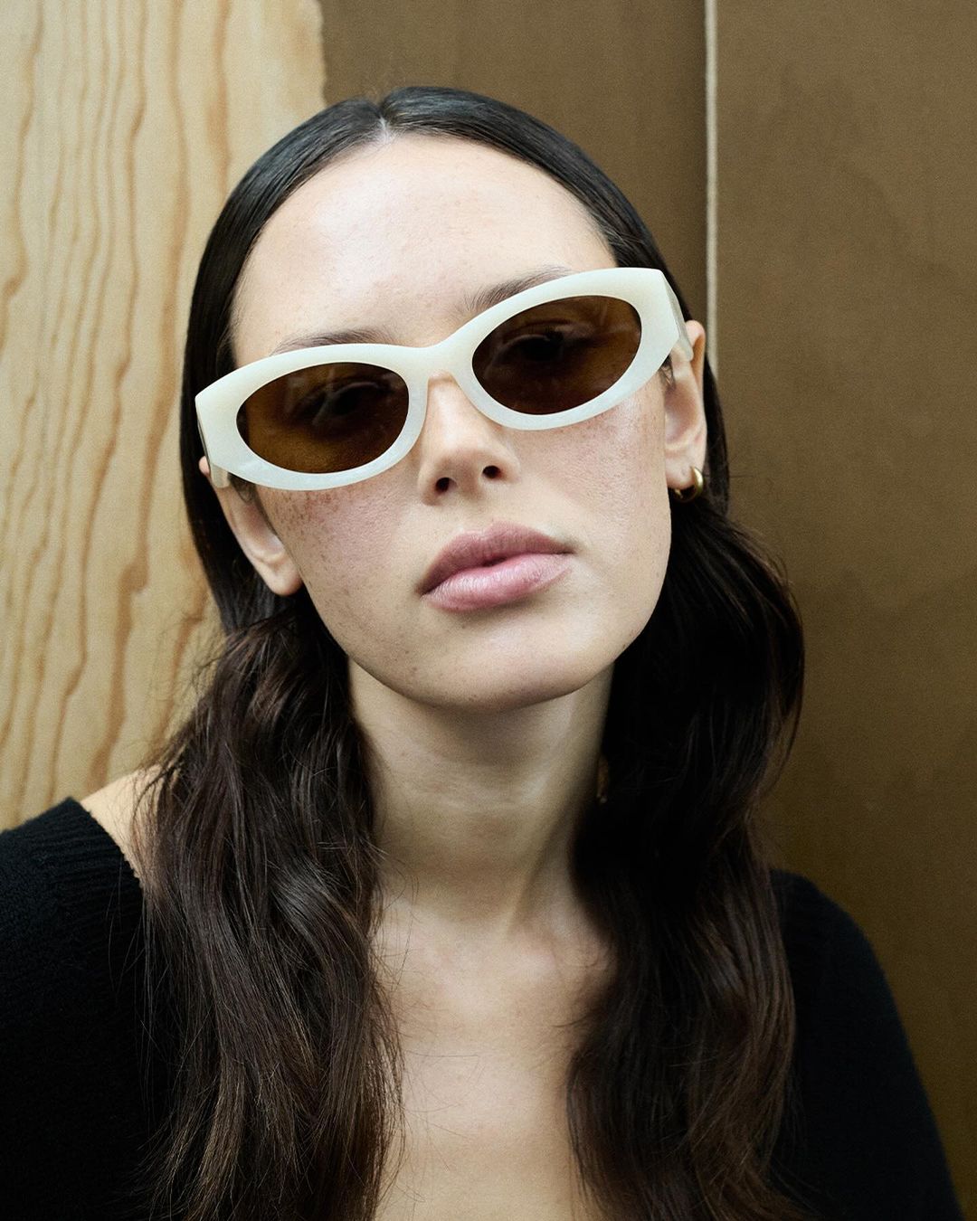 Brunette woman wearing off-white oval sunglasses with brown lenses, a black top and gold hoop earrings poses against a neutral background.