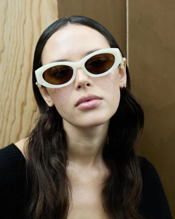 Brunette woman wearing off-white oval sunglasses with brown lenses, a black top and gold hoop earrings poses against a neutral background.
