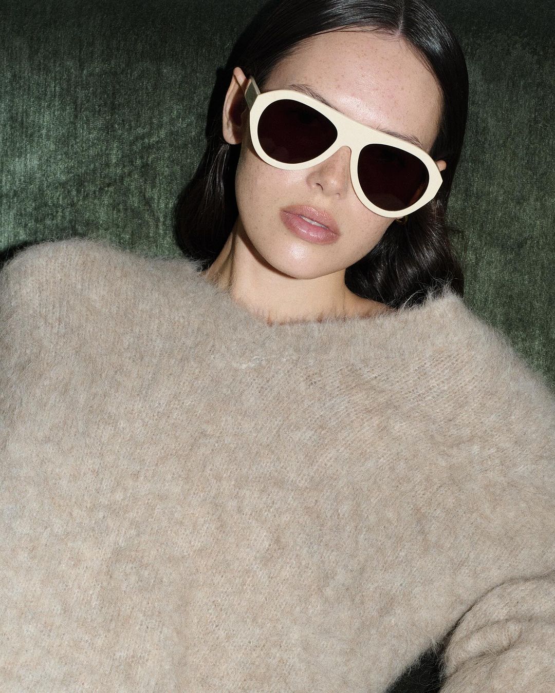 Close-up of an adult woman wearing sunglasses and a sweater, set against a muted green backdrop in an indoor setting. Fashion shot.