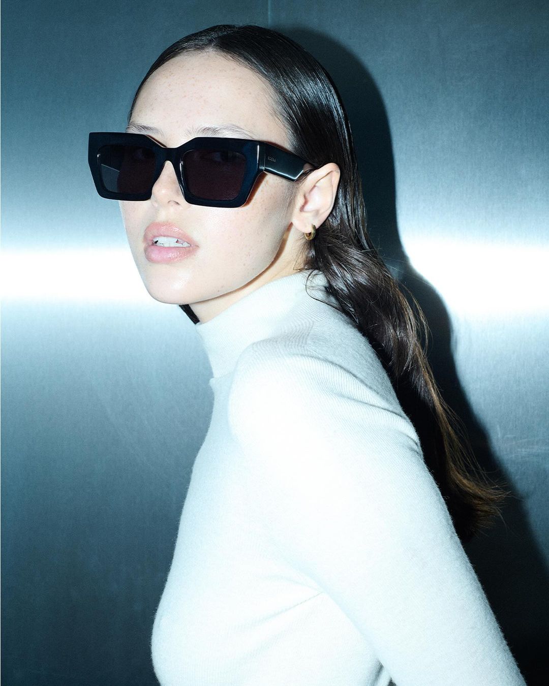 Model wearing dark sunglasses and a white turtleneck top against a metallic background for Edera Eyewear 2024 campaign.