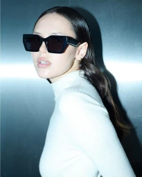 Model wearing dark sunglasses and a white turtleneck top against a metallic background for Edera Eyewear 2024 campaign.