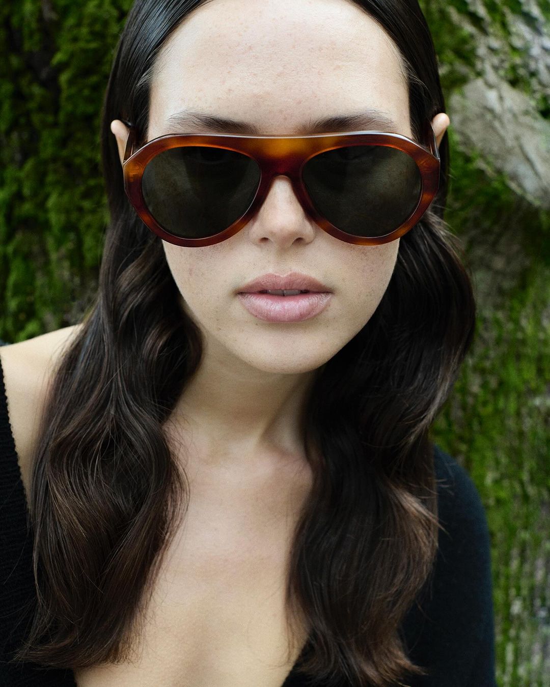 Brunette woman wearing tortoise shell sunglasses poses against a tree trunk in an outdoor environment, fashion, and beauty.