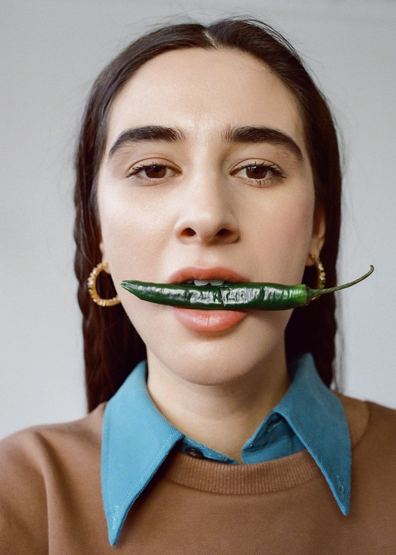 Portrait of a young woman with long dark hair and gold earrings holding a green chili pepper in her mouth against a neutral background.