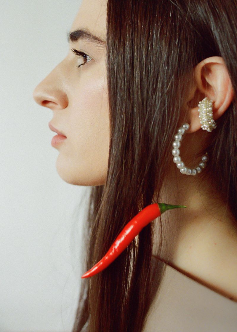 Profile of a woman with pearl earrings and a red chili pepper near her neck in a studio with a white backdrop for a Beauty product.