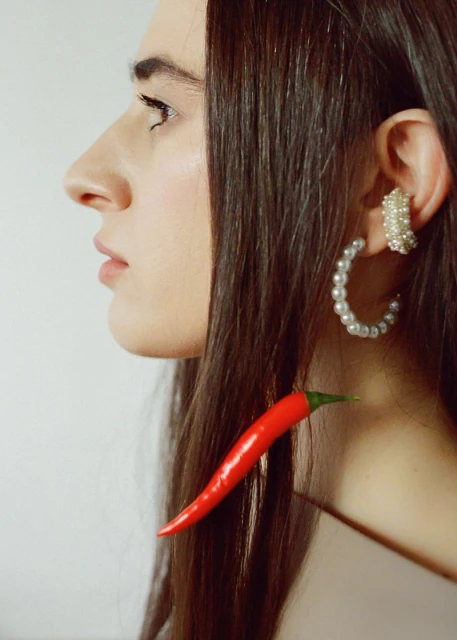 Profile of a woman with pearl earrings and a red chili pepper near her neck in a studio with a white backdrop for a Beauty product.