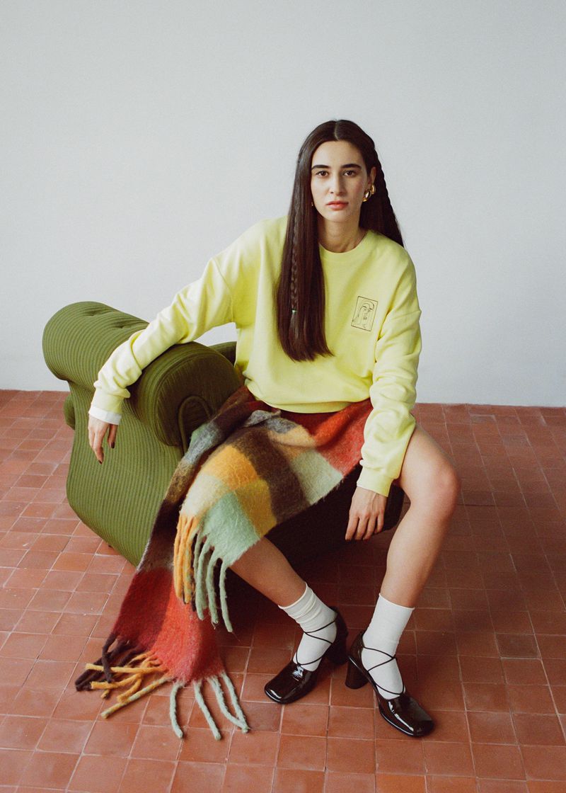 A woman sits in an olive chair wearing a yellow sweatshirt, short red skirt, black heels and a multi-colored, plaid scarf.