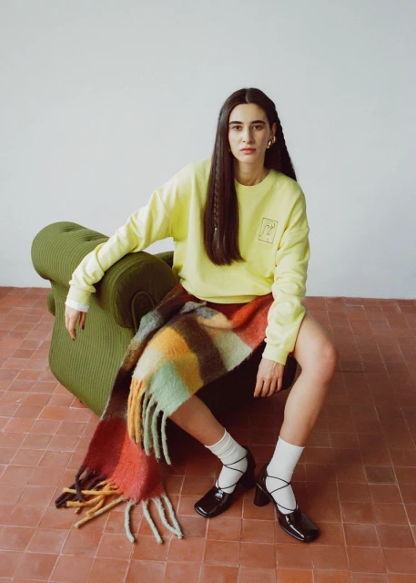 A woman sits in an olive chair wearing a yellow sweatshirt, short red skirt, black heels and a multi-colored, plaid scarf.