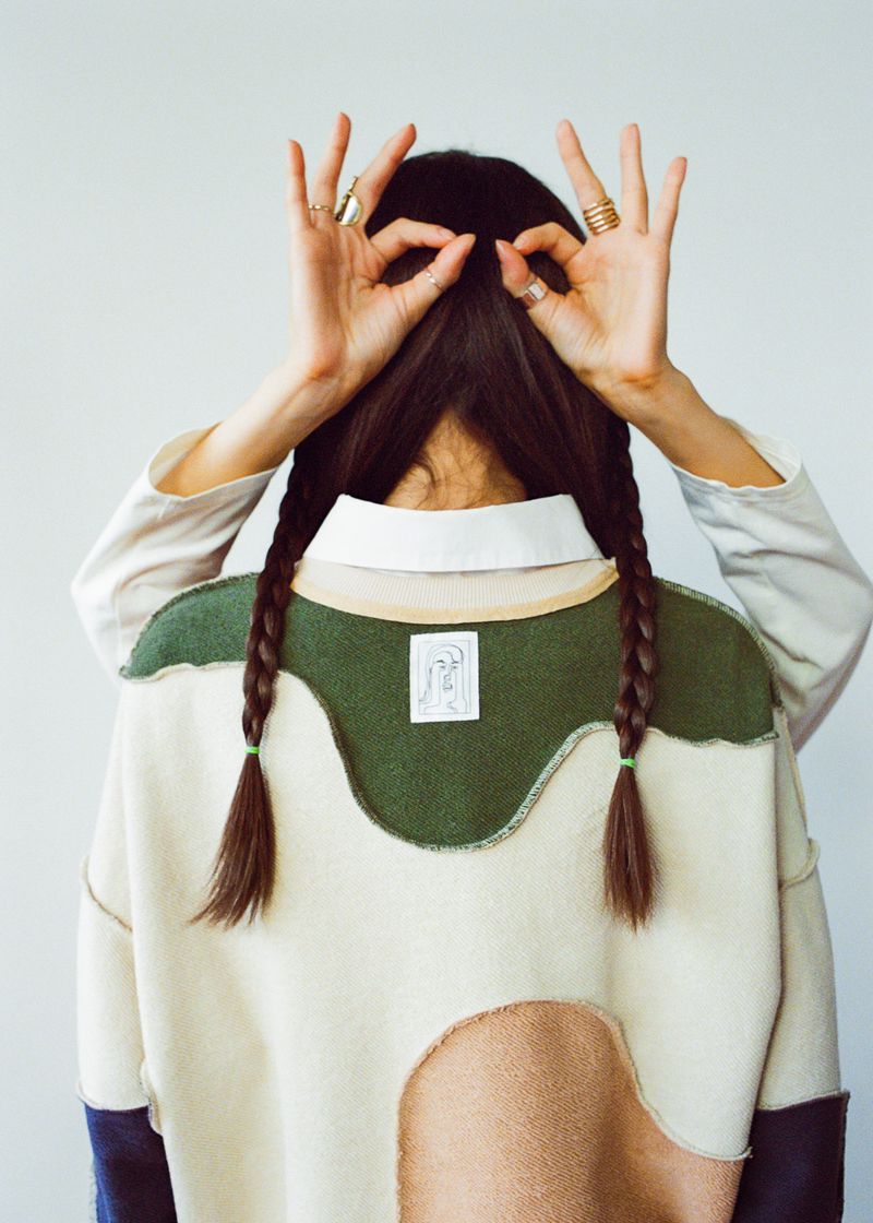 A woman with dark hair in braids makes an OK hand gesture over her head, wearing a sweater with geometric color blocks and a collared shirt.