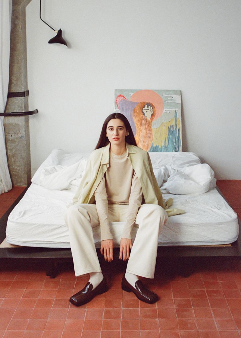 Adult female seated on a bed with a painting in the background, wearing cream-colored clothing and dark brown shoes in an indoor setting.