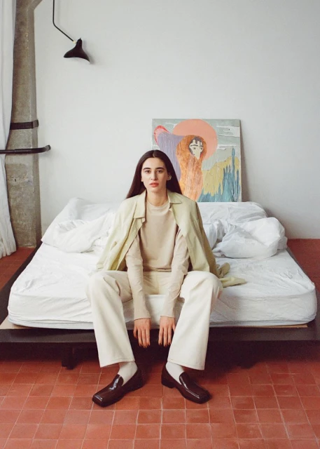 Adult female seated on a bed with a painting in the background, wearing cream-colored clothing and dark brown shoes in an indoor setting.