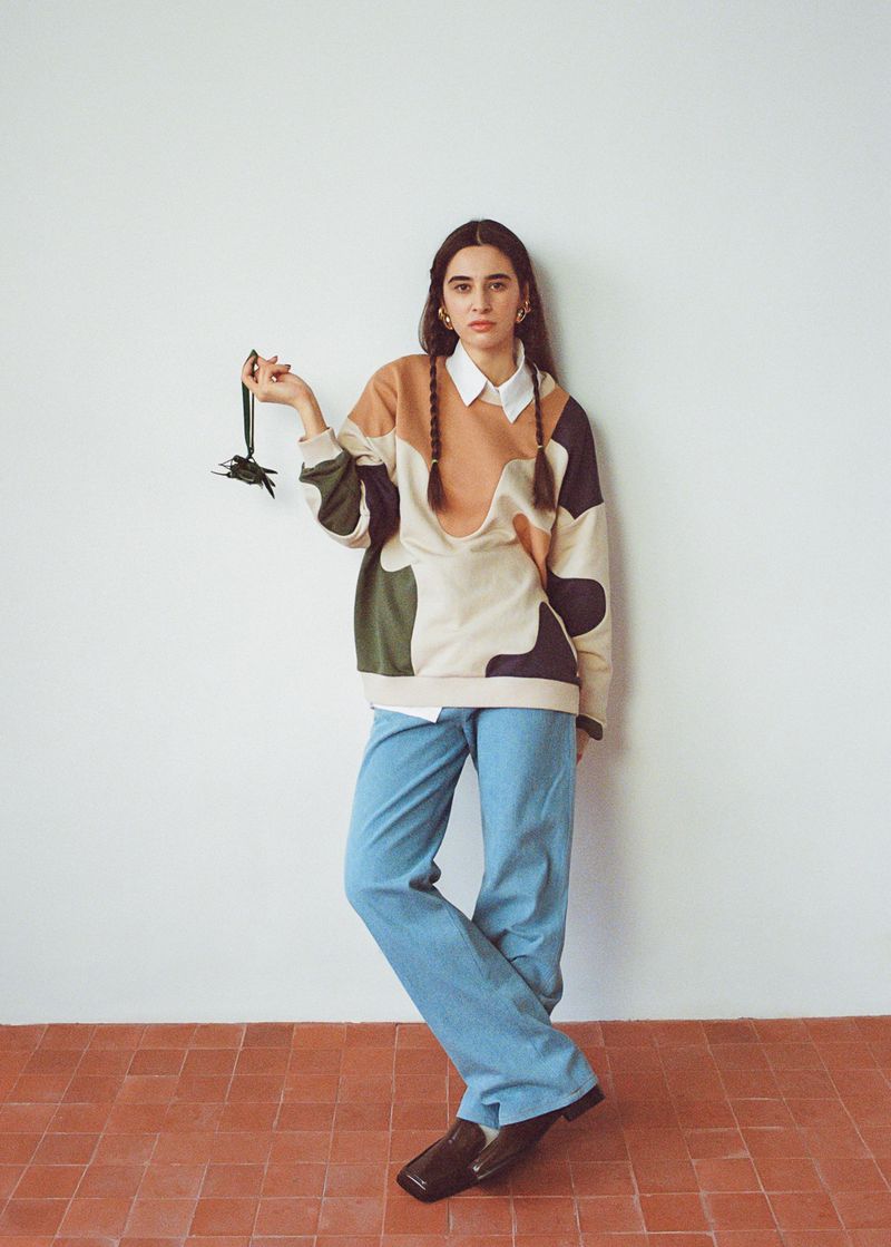 A young woman with braided hair stands in a studio wearing a color-blocked sweater and holding a black accessory, with a neutral backdrop.