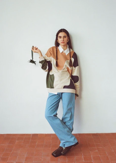 A young woman with braided hair stands in a studio wearing a color-blocked sweater and holding a black accessory, with a neutral backdrop.