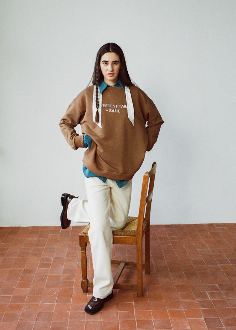 A woman poses in a brown sweatshirt, off-white pants, and black shoes, leaning against a wooden chair on a terracotta-tiled floor.