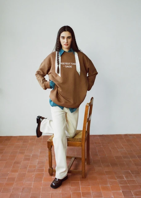 A woman poses in a brown sweatshirt, off-white pants, and black shoes, leaning against a wooden chair on a terracotta-tiled floor.