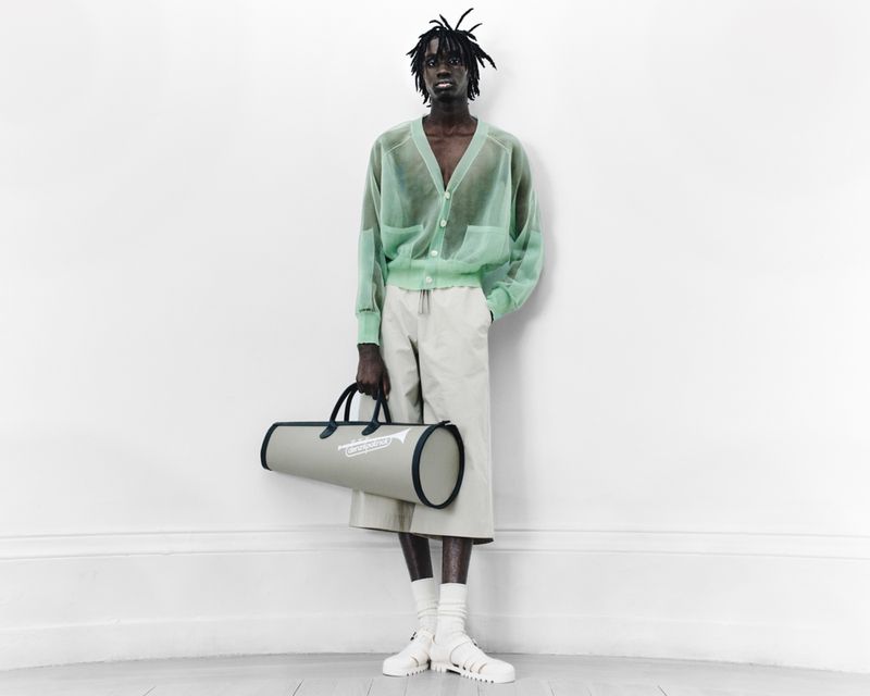 Male fashion model poses in a sheer green cardigan, cream shorts, and sandals, holding a cylindrical bag against a white wall.