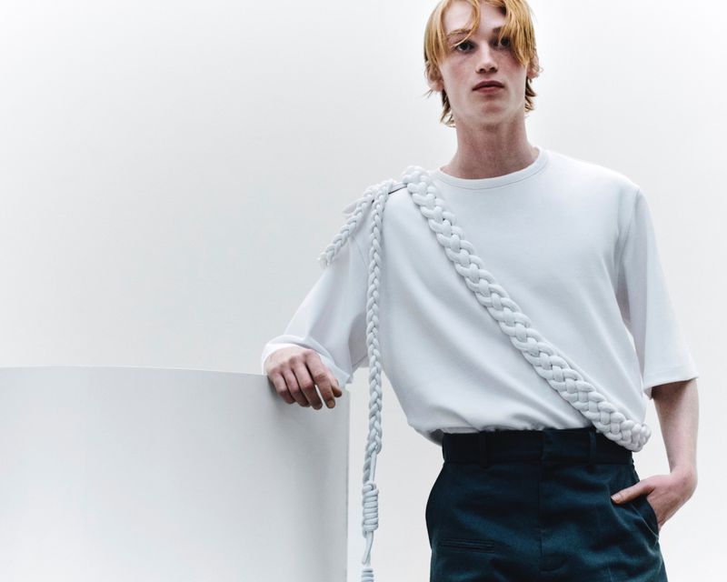 Man in white t-shirt and dark pants poses next to white wall, showcasing a braided white rope detail around his shoulder.