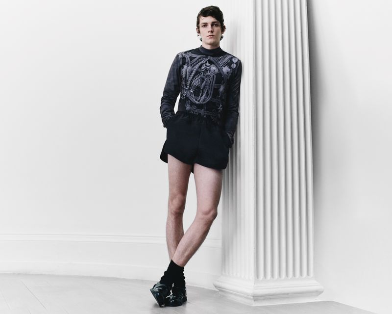 Young adult male fashion model poses in black long-sleeve top, black shorts, and black textured shoes against a white wall with pillar.
