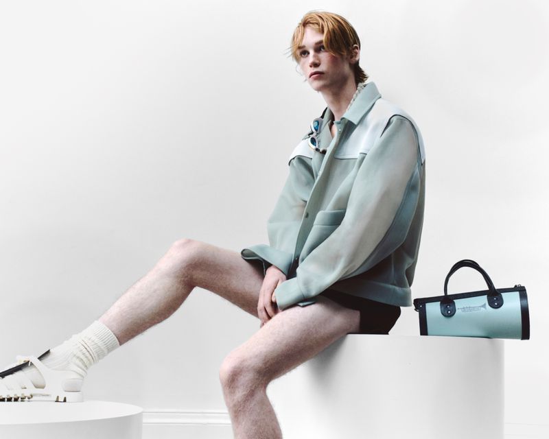 Man in stylish jacket and white sandals sitting against a plain background, accessorized with a necklace and a light blue bag.