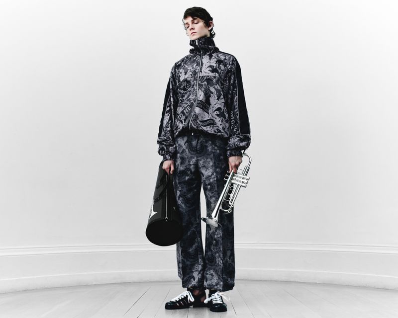 Fashionable adult male poses indoors wearing a patterned tracksuit, holding a trumpet and bag against a plain white wall backdrop.