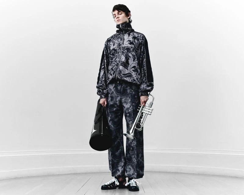 Fashionable adult male poses indoors wearing a patterned tracksuit, holding a trumpet and bag against a plain white wall backdrop.