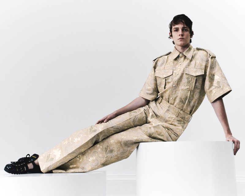 Man in patterned co-ord set poses against a bright white backdrop, seated on a white cylindrical prop in a studio setting, fashion shot.