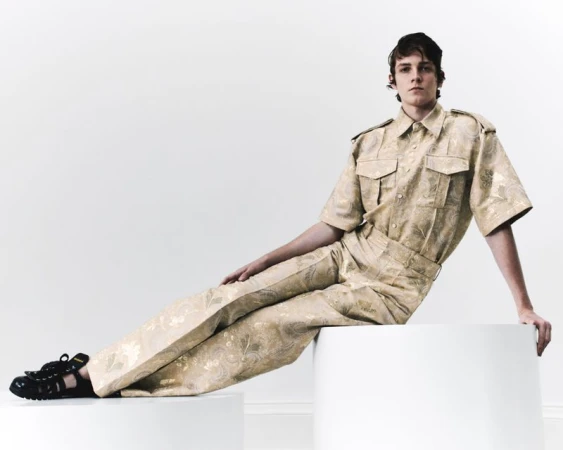 Man in patterned co-ord set poses against a bright white backdrop, seated on a white cylindrical prop in a studio setting, fashion shot.