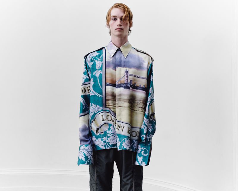 Fashion shot of a young adult man wearing a long-sleeved button-down shirt with a bridge graphic standing against a white backdrop.
