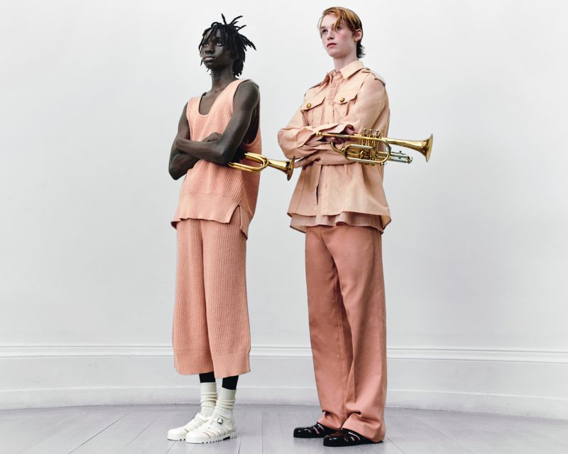 Two people in matching peach outfits, holding trumpets in a bright studio, showcasing fashion and musical instrument integration.