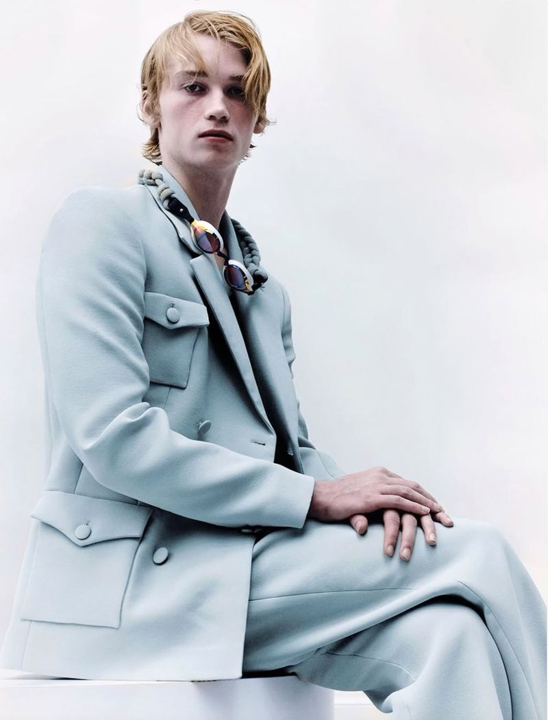 Portrait of a young male adult in a light blue suit sitting on a white surface, looking at the camera with a neutral facial expression.