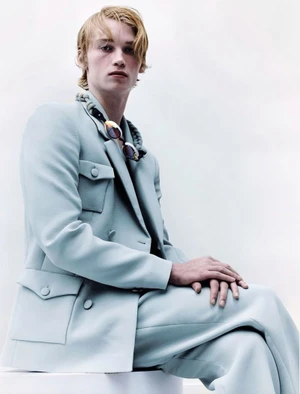 Blonde male model wearing a pale blue suit jacket and trousers seated against a bright white background