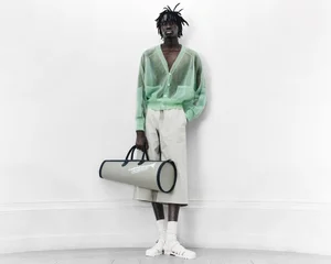 Model wearing a sheer mint green cardigan and grey shorts holding a cylindrical bag against a white wall.