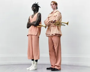 Two male models in peach-toned apparel holding brass trumpets against a light studio backdrop for Denzil Patrick S/S 2025 lookbook.