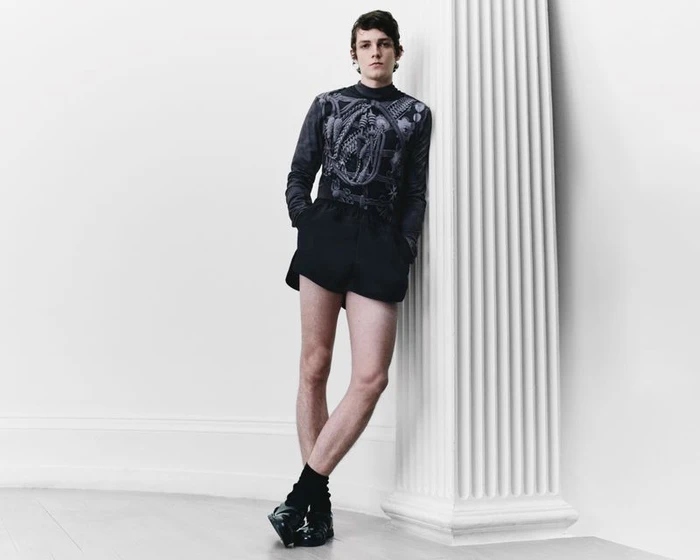Model leaning against a large white column wearing a patterned long-sleeve top and dark shorts for Denzil Patrick S/S 2025 Lookbook.