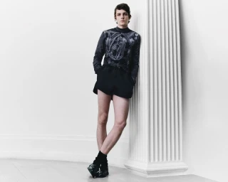 Model leaning against a large white column wearing a patterned long-sleeve top and dark shorts for Denzil Patrick S/S 2025 Lookbook.