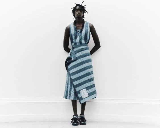 Model Babakar Ndiaye in a blue striped wrap garment against a neutral white wall for Denzil Patrick S/S 2025 Lookbook.