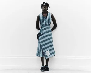 Model Babakar Ndiaye in a blue striped wrap garment against a neutral white wall for Denzil Patrick S/S 2025 Lookbook.