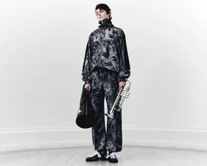 Model standing against a plain background holding a trumpet and a cylindrical case while wearing a patterned jacket and trousers.