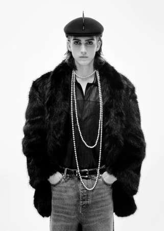 Black and white shot of a young adult wearing a faux fur coat, pearl necklaces, denim jeans, and beret against a white background.