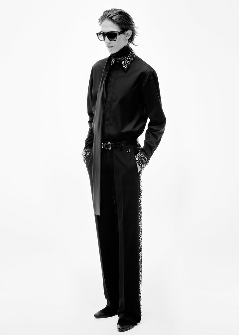 Black and white studio shot of a fashionable adult female wearing a black shirt, long scarf, bedazzled black pants, and black shoes.