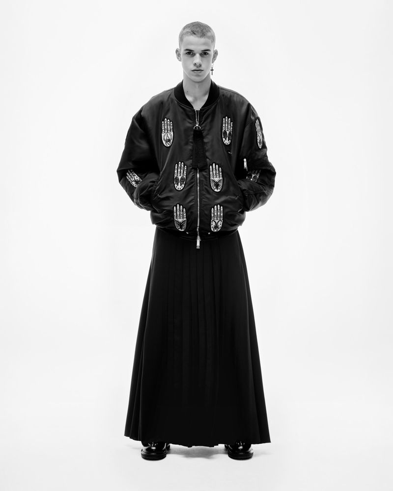 Monochrome studio portrait of a male fashion model wearing a black bomber jacket with hand designs and long pleated skirt against white.