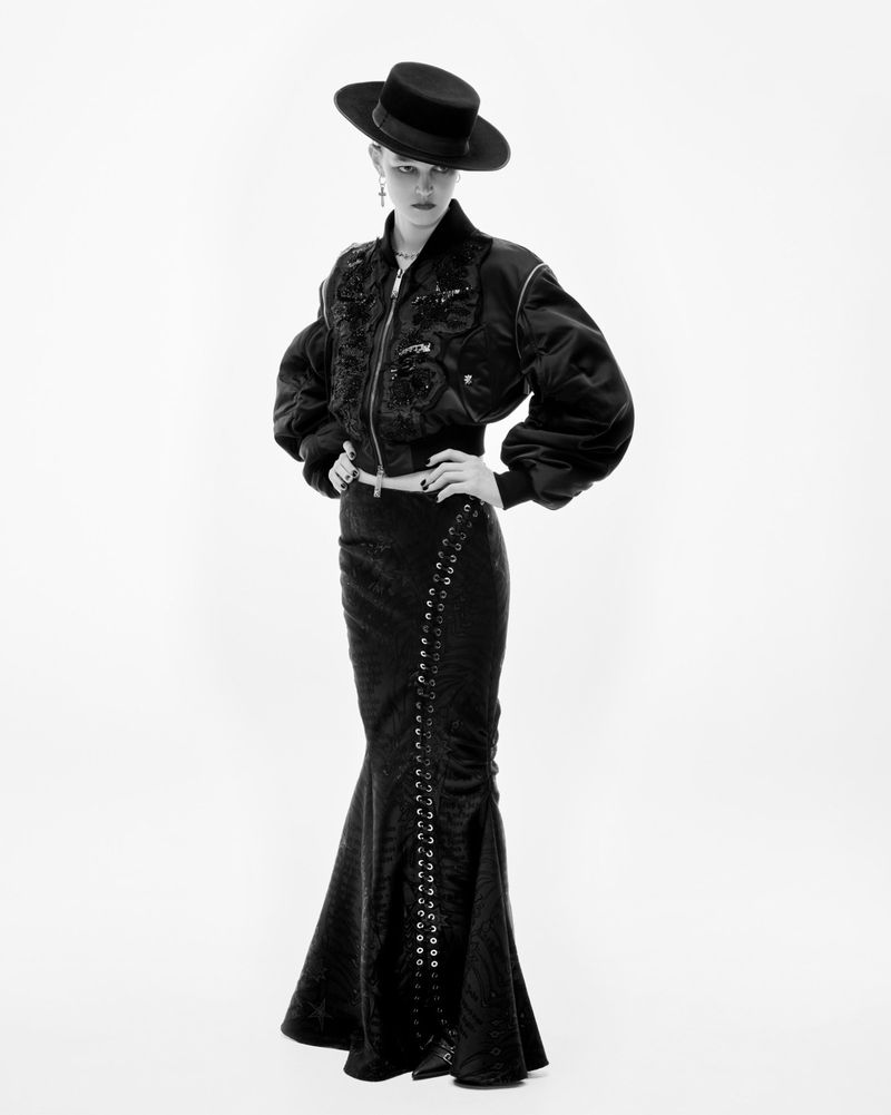 Monochrome fashion shot of a young woman in a cropped jacket, long skirt, and wide-brimmed hat, posing against a stark white backdrop.