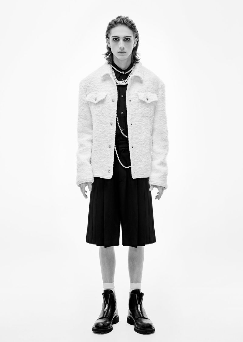 Monochrome fashion portrait of a male model wearing a white sherpa jacket, black shirt and shorts, pearl necklace, and black boots in a studio.