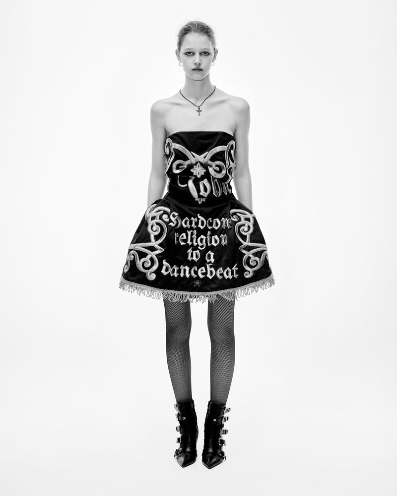 Fashion shot: Woman stands in a black strapless dress with white script and decoration, fishnet stockings, and black buckle boots on white.