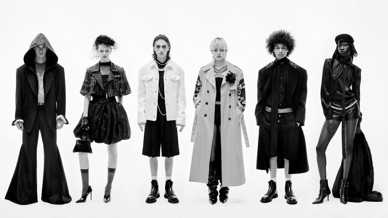 Black and white fashion shot of six people in a studio wearing designer clothing, including coats, dresses, and boots.