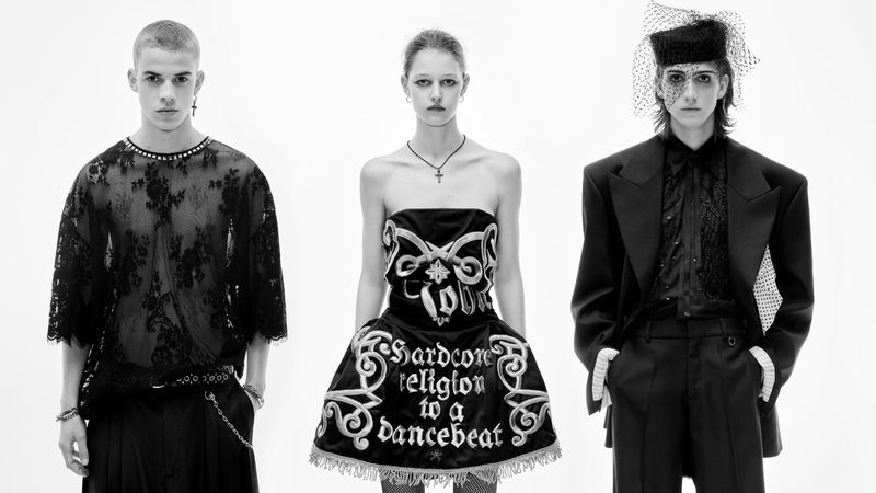 Three fashion models pose in a stark photo studio. The model in the center wears a black strapless dress with white embroidery and fringe.